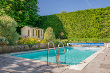 Outdoor swimming pool with a pale timber summerhouse set among mature trees and garden planting.